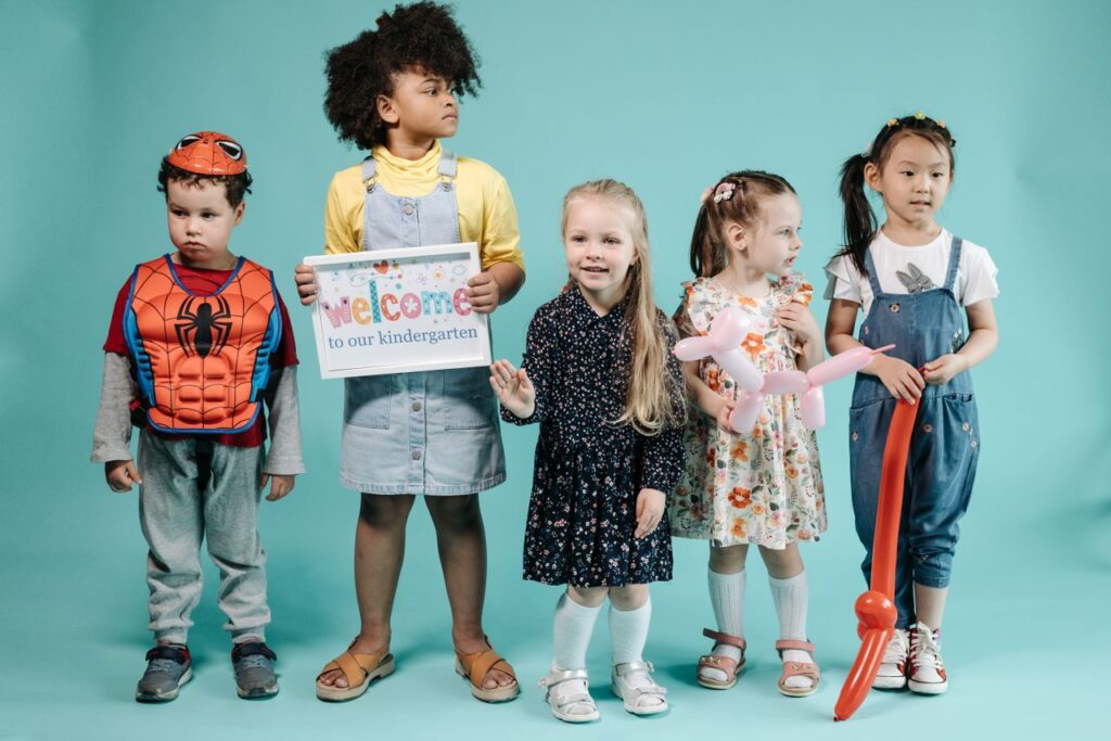 Five diverse children stand with a welcome to kindergarten sign in a studio setting.